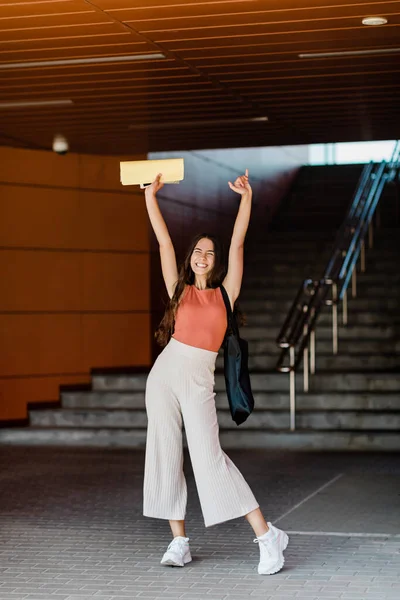 Seorang gadis yang ceria berdiri di dekat tangga, memegang sebuah folder di tangan yang ditinggikan dan tersenyum — Stok Foto