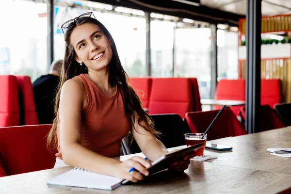 Uma menina pensativa está sentada em um café usando um tablet, fazendo anotações em um caderno. — Fotografia de Stock