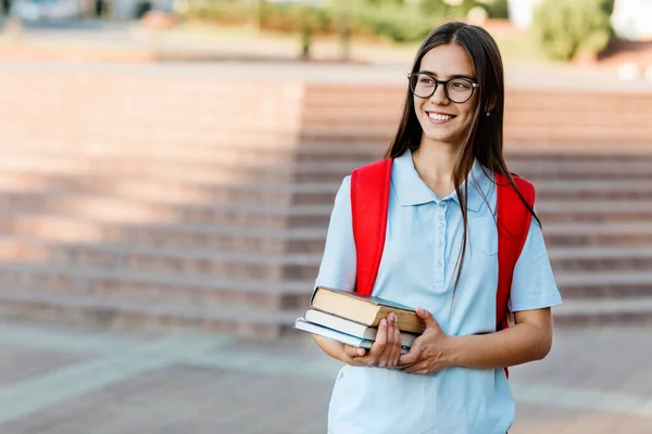 Una estudiante sonriente con gafas, libros y una mochila roja. Retrato de un estudiante en un fondo de la ciudad. El concepto de educación — Foto de Stock