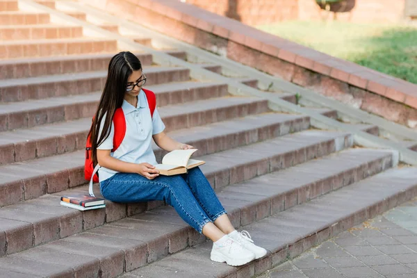 Una chica bonita estudiante en gafas, con una mochila está sentada en las escaleras cerca de la universidad, leyendo un libro, preparándose para conferencias o exámenes. Formación y educación —  Fotos de Stock