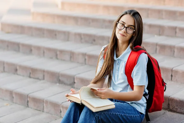 Una chica bonita estudiante en gafas, con una mochila está sentada en las escaleras cerca de la universidad, leyendo un libro, preparándose para conferencias o exámenes. Formación y educación —  Fotos de Stock