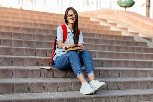 Una estudiante enfocada con gafas, sentada en las escaleras, escribe en su cuaderno. Preparación para una conferencia o examen — Foto de Stock