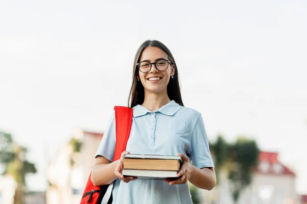 Una estudiante alegre con una mochila y libros, de pie y sonriendo en la calle — Foto de Stock