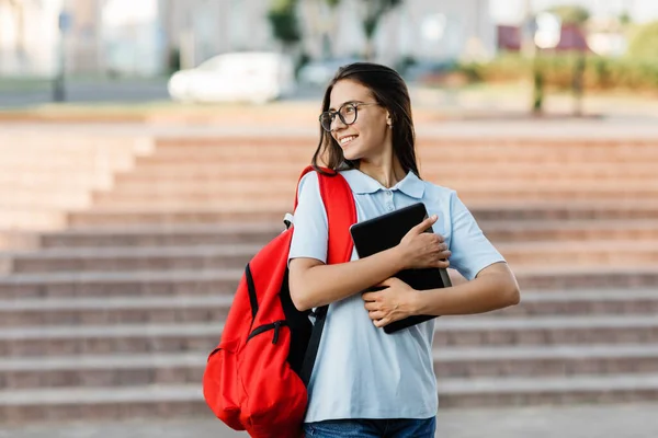 Uma estudante bonita e sorridente de óculos e com uma mochila vermelha, com um tablet nas mãos, fica ao lado das escadas e sorri — Fotografia de Stock