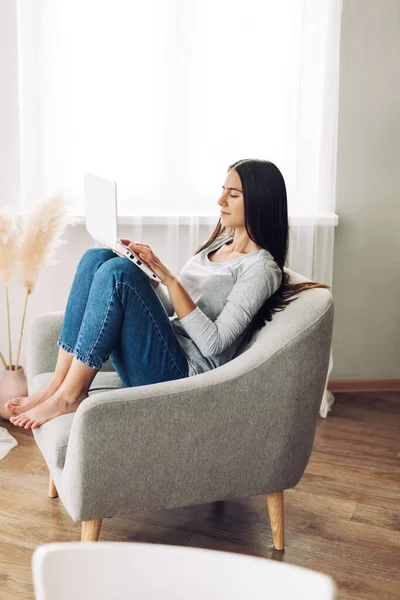 Uma menina bonita com um laptop está sentada em uma cadeira macia e confortável e trabalhando em um laptop, digitando texto. Tecnologias modernas, redes sociais — Fotografia de Stock