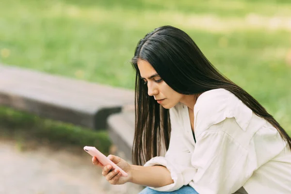Una chica escribe mensajes SMS o utiliza aplicaciones en un teléfono inteligente mientras está sentada en un banco en el parque — Foto de Stock