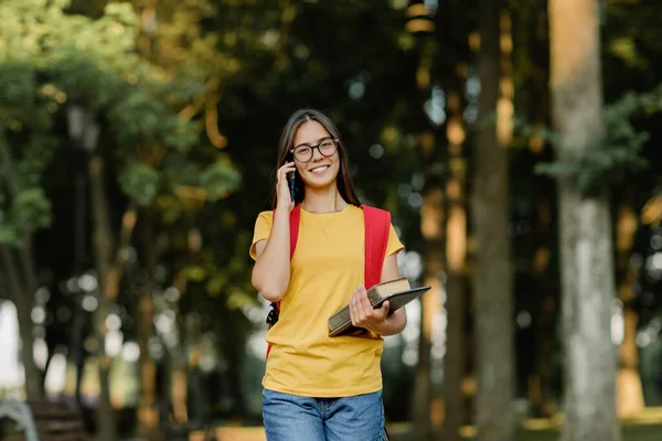 Portrét krásné brunetky s brýlemi a batohem mluvící po telefonu v parku — Stock fotografie
