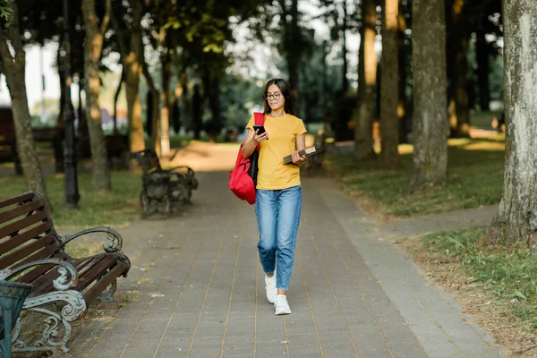Retrato de una joven estudiante caminando por el parque con una mochila, un libro y un teléfono en sus manos — Foto de Stock