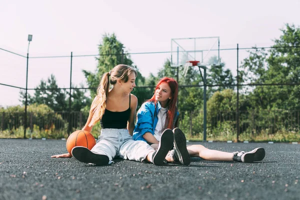 Две девушки в спортивной одежде и с баскетболом болтают, сидя на детской площадке. Спорт, конкуренция, дружба — стоковое фото