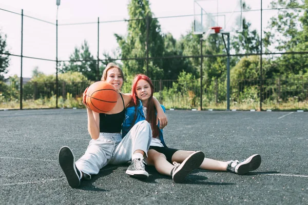 Dos chicas en ropa deportiva y con un baloncesto están charlando, sentadas en el patio de recreo. Deportes, competición, amistad —  Fotos de Stock