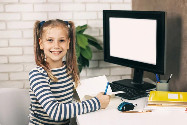 Блондинка с красивыми хвостиками сидит дома за компьютером, смотрит в камеру и улыбается. Ребенок учится на дому. Дистанционное обучение. — стоковое фото