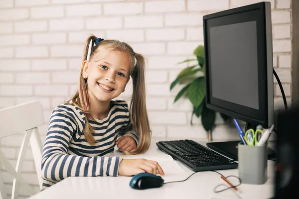 Una chica rubia con hermosas colas de caballo está sentada en una computadora en casa, mirando a la cámara y sonriendo. El niño aprende de casa. Aprendizaje a distancia online. —  Fotos de Stock