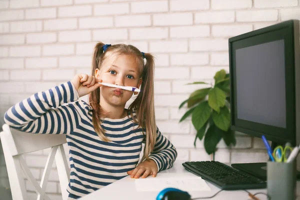 La colegiala se sienta en casa a la mesa en la computadora, juega con un lápiz, juega y ríe. Educación en línea. El niño aprende de casa. Aprendizaje a distancia online. — Foto de Stock