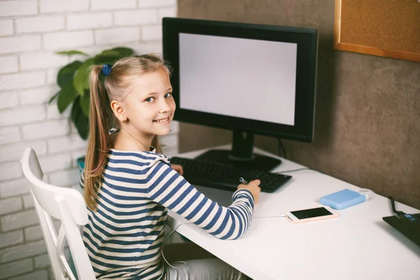 A schoolgirl in headphones sits at a table near a computer at home and does homework, looking at the camera and smiling. Online training. The child learns from home. Distance learning online. — Stock Photo, Image