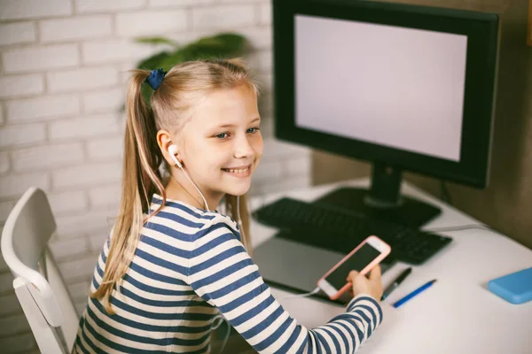 Una chica en auriculares con un teléfono en sus manos se sienta en casa en la mesa en el ordenador y hace su tarea. Educación en línea. El niño aprende de casa. Aprendizaje a distancia online. —  Fotos de Stock