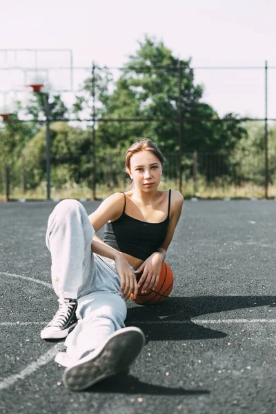 Retrato de uma menina encantadora sentada em um campo de esportes em um parque ou escola com um basquete depois de um jogo ou treino — Fotografia de Stock