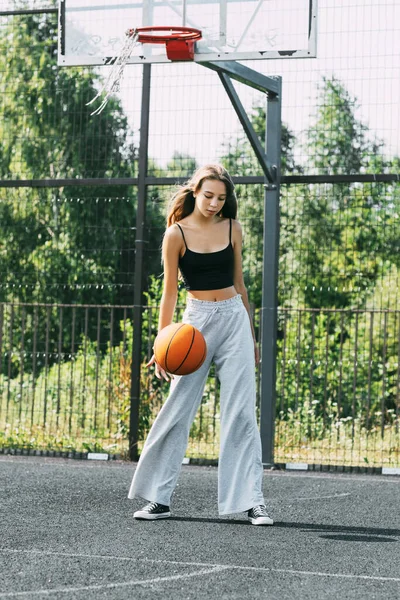 Una hermosa adolescente conduce un baloncesto en un campo de deportes, una chica aprende a jugar baloncesto —  Fotos de Stock