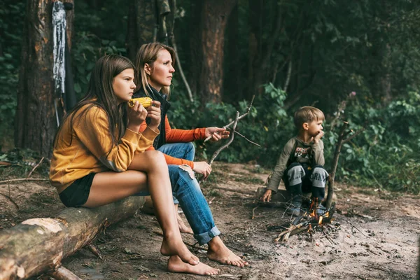 Uma jovem mãe com seus filhos está sentada na fogueira durante um feriado conjunto, uma garota está comendo milho. — Fotografia de Stock