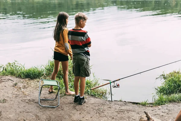 Ένας αδερφός και μια αδελφή στέκονται στην όχθη του ποταμού και ψαρεύουν κατά τη διάρκεια οικογενειακών διακοπών σε κάμπινγκ. Χρόνος μαζί, οικογενειακές διακοπές — Φωτογραφία Αρχείου