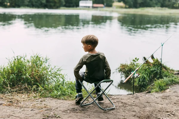 Το αγόρι κάθεται σε μια πτυσσόμενη καρέκλα στην όχθη μιας λίμνης ή ενός ποταμού. Αναψυχή, σαββατοκύριακα, τουρισμός. Πίσω όψη — Φωτογραφία Αρχείου