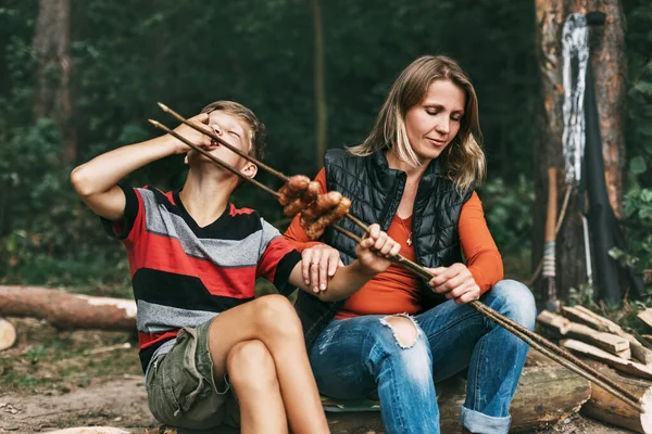 Une mère et son fils adolescent sont assis sur un arbre dans des campings et essaient des saucisses grillées cuites sur un feu — Photo