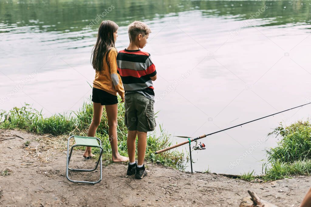 A brother and sister are standing on the river bank and fishing during a family vacation at a camping site. Time together, family vacation