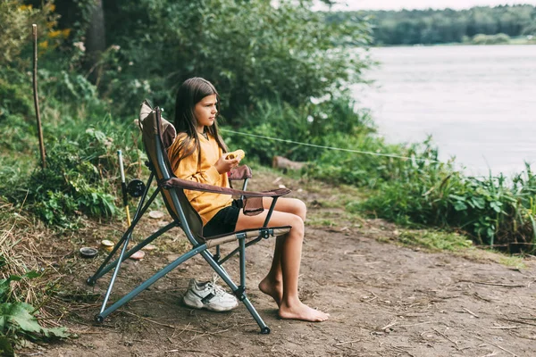Uma adolescente em uma camisola amarela brilhante está sentada em uma cadeira de acampamento na margem do lago e comendo milho — Fotografia de Stock