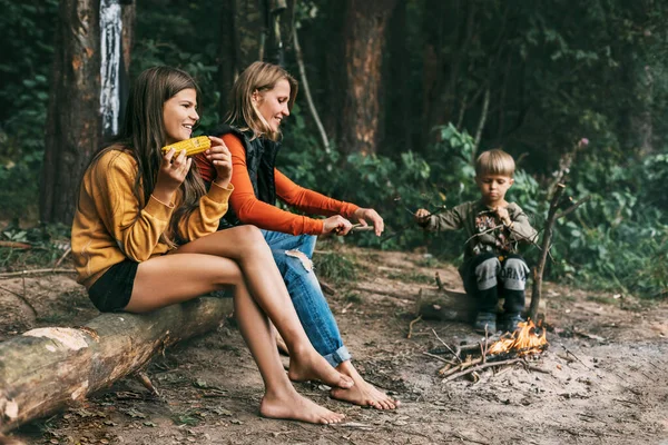 Una joven madre con sus hijos está sentada junto a la fogata durante unas vacaciones conjuntas, una niña está comiendo maíz —  Fotos de Stock