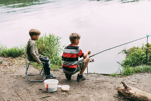 Two brothers sit on the shore of a lake or river and fish with fishing rods. The brothers are waiting for their catch