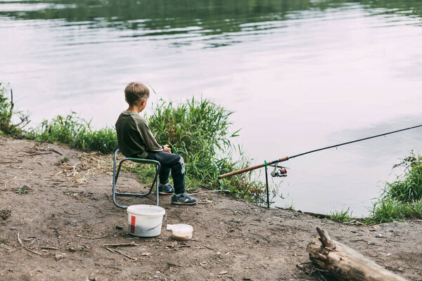 A boy is sitting on the shore of a lake or river with a fishing rod during a family vacation, a child is waiting for his catch