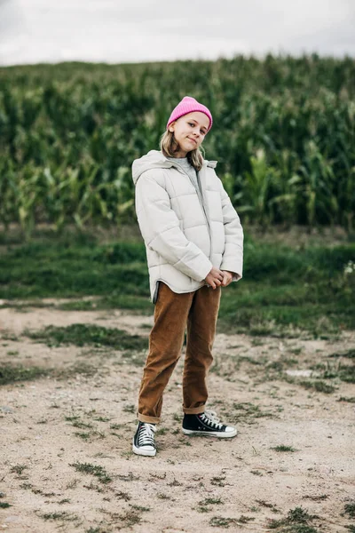 Vor dem Hintergrund eines Maisfeldes steht ein Mädchen im Teenageralter mit stylischem Image und rosa Mütze. — Stockfoto