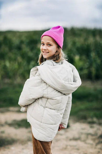 Fröhlich schönes Teenager-Mädchen in gelbem Regenmantel und rosa Hut tanzt auf dem Hintergrund eines Maisfeldes — Stockfoto