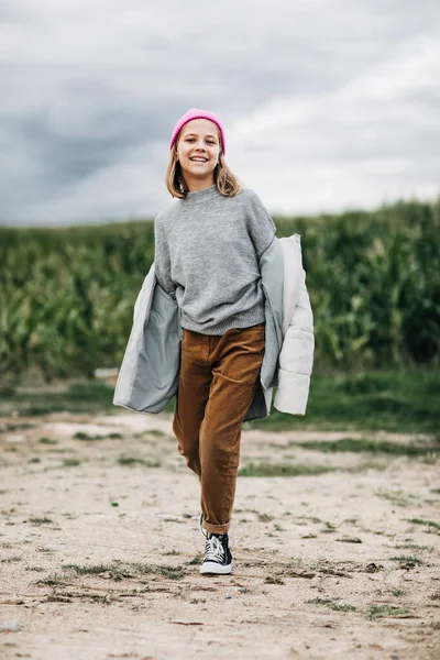 Menina adolescente bonita alegre em capa de chuva amarela e chapéu rosa confiantemente caminha no campo de milho — Fotografia de Stock