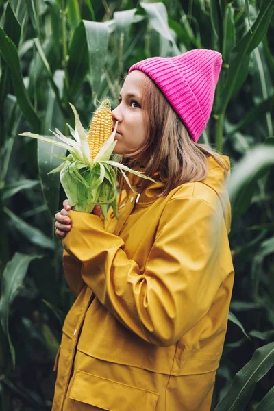 Engraçado menina vestida com uma capa de chuva amarela e um boné rosa quente estraga e morde milho em um milheiral — Fotografia de Stock