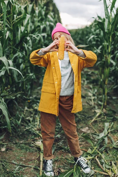 Lustiges kleines Mädchen in einem gelben Regenmantel und einer knallrosa Mütze verwöhnt und beißt Mais in einem Maisfeld — Stockfoto