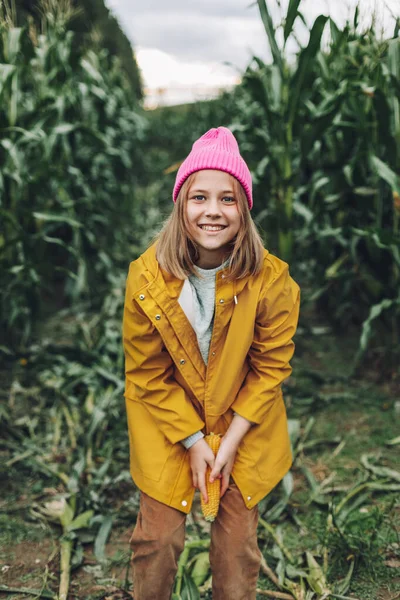 Niña divertida vestida con un impermeable amarillo y una gorra rosa caliente echa a perder y muerde maíz en un maizal —  Fotos de Stock