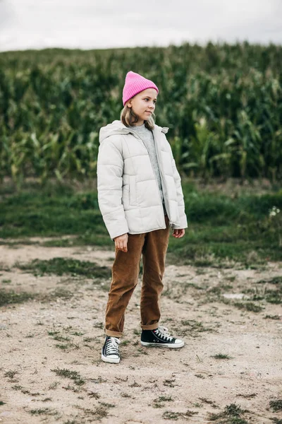 Vor dem Hintergrund eines Maisfeldes steht ein Mädchen im Teenageralter mit stylischem Image und rosa Mütze. — Stockfoto