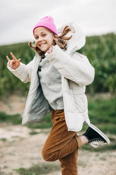 Alegre hermosa adolescente en un impermeable amarillo y sombrero rosa saltando sobre el fondo de un campo de maíz —  Fotos de Stock