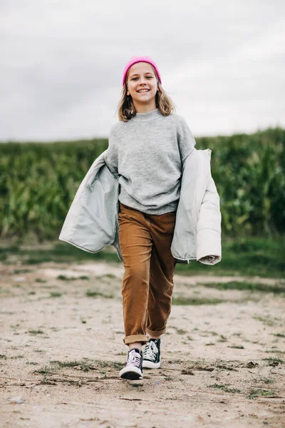 Vrolijk mooi tiener meisje in gele regenjas en roze hoed loopt vol vertrouwen in het maïsveld — Stockfoto