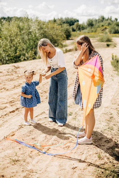 Uma menina pequena bonito junto com sua mãe e irmã estão preparando um papagaio para o lançamento durante umas férias fora da cidade — Fotografia de Stock