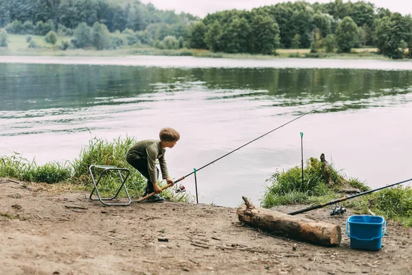 Ένα αγόρι στέκεται στην όχθη μιας λίμνης ή ενός ποταμού με ένα καλάμι ψαρέματος κατά τη διάρκεια των οικογενειακών διακοπών, ένα παιδί περιμένει την σύλληψή του. — Φωτογραφία Αρχείου