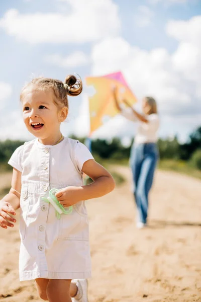 Uma menina em um vestido branco está correndo ao longo da estrada com uma corda de um papagaio na mão, uma menina está voando um papagaio com sua mãe — Fotografia de Stock