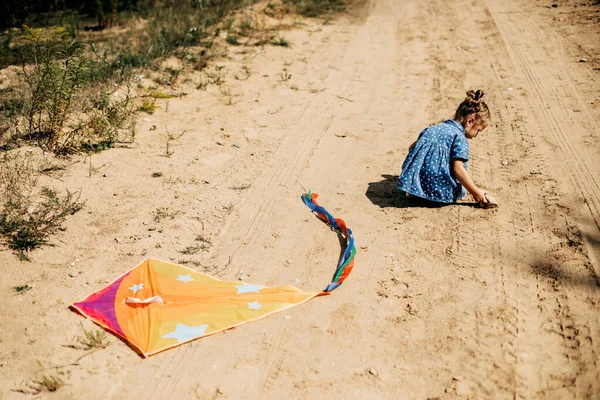 Vista trasera de un bebé en un vestido azul y una cometa acostada en un camino arenoso —  Fotos de Stock