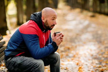 Yakışıklı sakallı bir adam sonbahar parkında bankta oturuyor ve sıcak çay içiyor.
