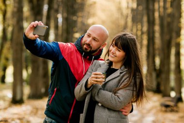 Birbirine aşık genç bir çift, sonbahar parkında yürürken telefonlarında fotoğraf çekiyor. Aşk ve ilişkiler kavramı