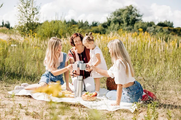 Drie generaties vrouwen van dezelfde familie drinken thee uit mokken tijdens een picknick in het park of op het gazon — Stockfoto