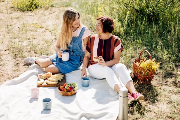 Eine lächelnde Mutter und ihre Tochter picknicken mit Obst und Gebäck auf dem Rasen. Einheit, gemeinsame Zeit, Elternschaft — Stockfoto