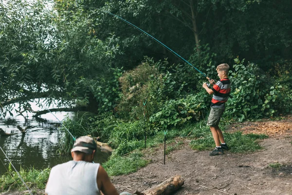 Ένας έφηβος με καλάμι ψαρέματος στέκεται σε μια βάρκα. ένα παιδί πιάνει ψάρια μόνο του στην όχθη μιας λίμνης ή ενός ποταμού και πιάνει ψάρια ενώ χαλαρώνει με την οικογένειά του — Φωτογραφία Αρχείου