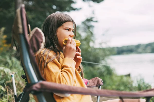 Μια έφηβη με ένα φωτεινό κίτρινο πουλόβερ κάθεται σε μια καρέκλα κάμπινγκ στην όχθη της λίμνης και τρώει καλαμπόκι. — Φωτογραφία Αρχείου