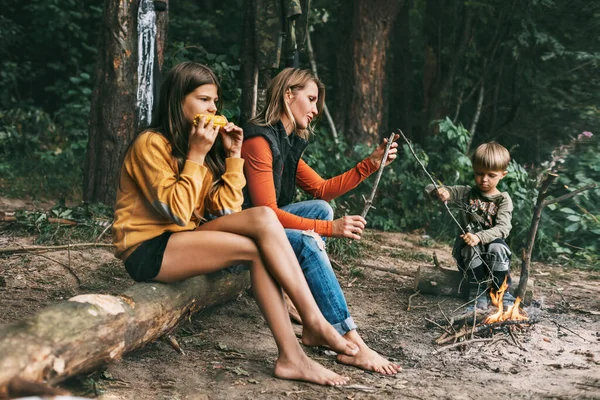 Μια νεαρή μητέρα με τα παιδιά της κάθεται δίπλα στη φωτιά κατά τη διάρκεια μιας κοινής γιορτής, ένα κορίτσι τρώει καλαμπόκι — Φωτογραφία Αρχείου
